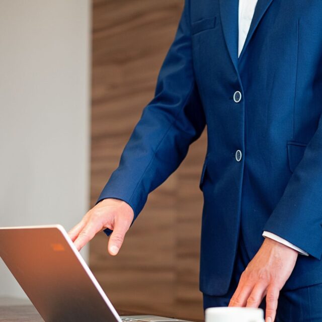 Business professional in a suit using a laptop on a desk in an office setting.